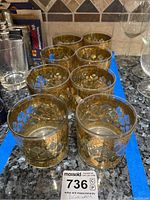 Set of eight vintage-style, short clear glasses with gold floral and geometric design embellishments, shown in two rows on a kitchen counter.