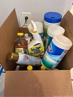 Top and side view of a cardboard box containing multiple cleaning products, showing product labels and container types.