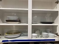 Kitchen cabinet shelf with stacks of white plates, clear glass plates, several white bowls and small glass bowls, vintage white mugs, and black plates.