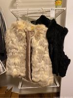 Two rabbit fur coats hanging on a white rack: one light beige and the other black, showing entire coats with fur visible.