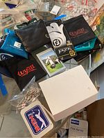 Various golf accessories arranged on table including wallets, gloves, golf bags with logo, Sky Caddie GPS device, box of golf balls and key chains.