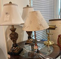 Four lamps on glass and wood table near window. Includes two ornate table lamps, one smaller textured shade lamp, and one brass desk lamp.