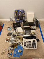 Wide view showing all items in the lot including walkie talkies in packaging, calculators, adding machines, tape recorder, locks, and charger