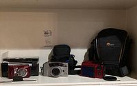 Shelf with Nikon Coolpix L20, Canon PowerShot Z85, red camera, and Lowepro camera carrying case