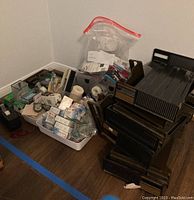 Photo showing grouped various office supplies including black plastic stackable file trays with wood accent handles, tape rolls, pens, small storage bins, staplers, and paper clips.