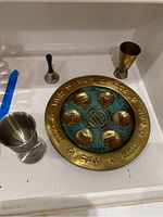 Large brass and turquoise Seder plate with Hebrew text and Star of David motifs, surrounded by the two chalices and the bell on a white surface.