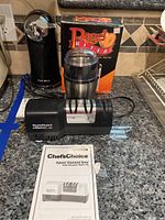 Overview of the kitchen countertop showing the collection of small kitchen appliances: bagel slicer in box, coffee grinder, electric can opener, and Chef'sChoice knife sharpener with manual in front.
