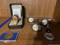 View of the lot showing four wristwatches—including gold-tone Valero Texas Open Fossil watch—and a fob watch, plus the NASA Project Apollo collectible coin in a display case.