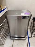 Closed rectangular stainless steel trash bin showing front view with pedal base and stickers on top.