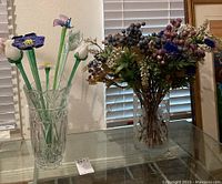 Two glass vases displayed side by side on a glass table. One vase is tall and cut with geometric patterns; the other holds an arrangement of faux glass flowers with long stems and colorful heads.