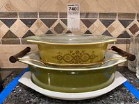 Two stacked Pyrex serving casserole dishes on a kitchen counter, showing the larger olive green casserole with a clear lid inside a metal holder with wooden handles, and the smaller white dish below.