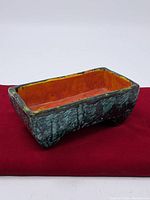 Side angle photo showing mottled turquoise textured exterior and orange interior of rectangular ceramic vase on red fabric.
