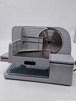 Front angle view showing the stainless steel serrated blade, food carriage with guard, and base with clear plastic tray.