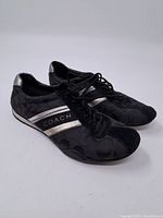 Front and side view of Coach shoes showing black fabric with Coach signature pattern and silver stripes with 'COACH' text.