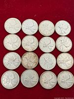 Top view of sixteen Canadian silver quarters arranged in four rows on a red background, showing reverse side with caribou design.