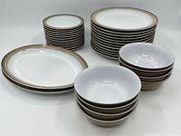 Front view of complete lot showing stacked bowls, plates, and platters in white ceramic with brown rim.