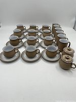 Overall view of 16 cups with saucers, 4 tall mugs and 2 lidded bowls on white background