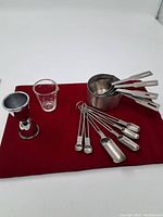 Full lot of measuring utensils arranged on a red cloth showing metal nested measuring cups, a glass measuring cup, a metal jigger, and measuring spoons and scoops.