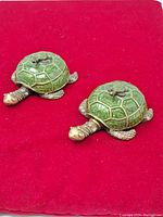 Two metal turtle-shaped trinket dishes with green enameled shells on a red cloth background.