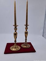 Pair of vintage solid brass candle holders standing on a red cloth, with tall tapered candles inserted.