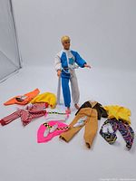 Full view of Ken doll in jumpsuit with outfits and shoes around him.
