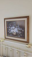 Photo showing the full framed floral art print hanging on a wall above a metal bed frame.
