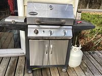 Front view of stainless steel Patio Chef BBQ grill with side shelves and cabinet, showing propane cylinder to the right.