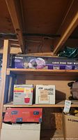 Photo of wooden shelf with boxed Spalding Cabin Dome Tent and various boxed lanterns below it, along with a piece of luggage visible beneath shelf.