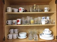 Shelf with various CorningWare mugs and plates with two different patterns, also clear glasses and Canadian-themed mugs.