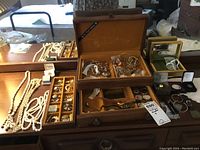 Wide shot of jewellery box on wooden table with various necklaces, bracelets, watches, pins, and jewelry items arranged outside and inside the box.