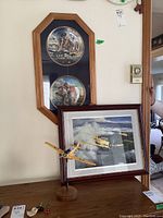Framed collector plates mounted in light wood octagonal frame with dark blue mat. Framed Harvard plane print in dark wood frame with double mat. Yellow Harvard model plane on wooden stand in front on table.