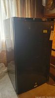 Black GE mini fridge shown from front near window, door closed.