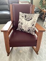 Front angle of wooden rocking chair upholstered in maroon vinyl with a decorative pillow, showing armrests, seat, and backrest.