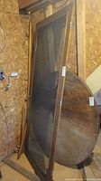 Wood-framed basement screen door leaning upright with visible wood grain, metal screen mesh, and attached metal hardware including handle, latch, and hinges.