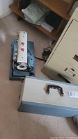 Sewing machine on floor with closed carrying case nearby, power cord visible.
