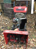 Front-on view showing red housing, black auger blades, pneumatic tires, handlebar and chute support frames.