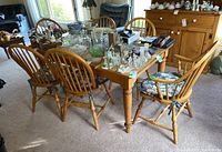 Full view of wood dining table and six spindle back chairs arranged around it with various decorative glass and ceramic items on the tabletop.