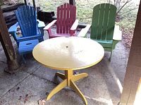 Photo showing round yellow table with three wooden Adirondack chairs in blue, pink, and green arranged around it outside.