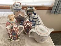 Full view of seven decorative ceramic teapots arranged on a white lace tablecloth including cottage shapes, a white and blue floral, and a rabbit design.
