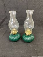 Front view of two green glass oil lamps with clear chimneys and brass tops on a black backdrop.