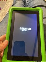 Tablet powered on displaying Amazon boot screen, held in hand, showing the green protective kid-proof case.