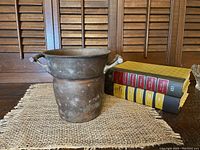 Small round copper pail bucket placed next to books on a woven mat with wooden background. Shows full front view of the bucket with bamboo style handle.