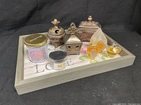 Wooden tray holding three bronze colored containers, three perfume bottles, and two candles.