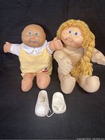 Two Cabbage Patch dolls side by side on black background with two white doll shoes in front.