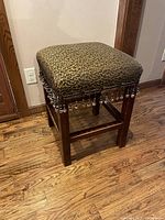 Front angle view of square animal print upholstered counter height barstool on wooden floors, showing beaded trim and dark wooden legs