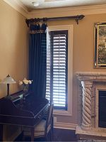 Curtain panels hanging in a room over window with plantation shutters, showing navy fabric with decorative gold and burgundy trim at the top and sides.