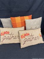 Five fall throw pillows displayed against dark cloth backdrop: two rectangular burlap pillows with 'gather' embroidery and autumn jar designs, two plain square burlap pillows with button details, one square orange and yellow checkered flannel pillow.