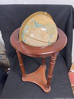 Front and side angles of the globe mounted on a dark brown wooden stand with three turned legs and a triangular base on a black cloth background showing the overall condition and height.