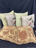Six throw pillows including four green burlap-style pillows with buttons, two beige burlap pillows with bird prints, and two gold floral velour pillows with fringed edges arranged on a dark cloth background.