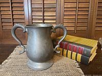Front view of the heavy pewter double handle tankard standing on a textured surface with books in the background
